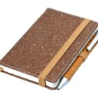 A closed brown notebook with a textured cover, featuring an attached elastic band. A wooden pen is held in a loop on the side. The notebook has rounded corners and a ribbon bookmark peeks out.