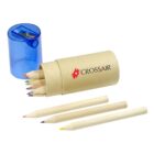 A small cardboard tube with colored pencils, labeled Crossair, lies on its side. Blue sharpener cap is removed, placed next to the tube. Three pencils are outside: one yellow, one red, and one purple.