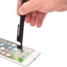 A hand uses a black stylus to interact with a smartphone displaying various app icons on its screen, placed on a white surface.
