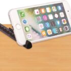 A smartphone with a light-colored case is propped up at an angle on a wooden surface using a triangular black stand that resembles a ruler. The screen displays app icons.