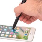 A hand using a black stylus to tap on a smartphone screen displaying various app icons. The phone is lying on a flat surface, and the hand is coming in from the top right corner of the image.