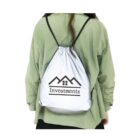 A person wearing a light green jacket and black skirt is seen from the back, carrying a white drawstring backpack. The backpack features a logo with stylized rooftops and the word Investments.