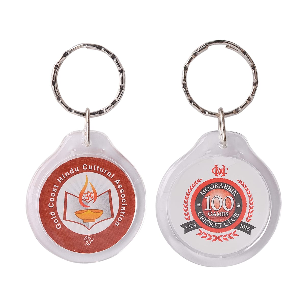 Two round keychains with metal rings. The left keychain features the Gold Coast Hindu Cultural Association logo with a lamp design. The right keychain displays the Moorabbin Cricket Clubs 100th games emblem with red and black accents.