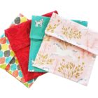 Four colorful fabric pouches are stacked, each with different patterns: multicolored leaves, red with small berries, teal with white animals, and pink with unicorns and trees.