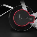A circular Fantasy wireless charger with a red and black design sits on a dark surface, next to a silver smartphone and a laptop.