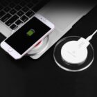 A smartphone with a charging icon on the screen rests on a round wireless charger connected to a laptop. The setup is on a dark surface, and the charger has a transparent outer ring.