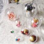 Clear globes on a lace surface, each containing different items: a pink rose, small plush toys, assorted colorful candies, and a jar with purple flowers.