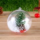 A clear ornament with a small artificial pine branch and a tiny pine cone inside, resting on a layer of white foam beads resembling snow, placed on a wooden surface with blurred background details.
