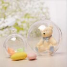 Two clear plastic spheres contain a small teddy bear with a blue bow, and pastel-colored candies. The spheres are placed on a light surface with a blurred floral background.