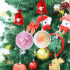 Close-up of a Christmas tree adorned with various ornaments, including two round transparent baubles filled with pink and orange materials, red velvet bags with Santa designs, a white stuffed bear, a bell, and a gold and red bauble.