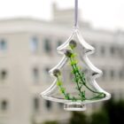 A transparent Christmas tree-shaped ornament hangs outdoors, containing green sprigs and small buds. In the blurred background, theres a multi-story building with several windows.
