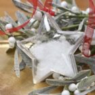A star-shaped clear ornament filled with white powder, resembling snow, hangs by a red ribbon. Its placed on silver leaves adorned with white berries, all set on a golden surface.