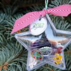 A clear star-shaped ornament filled with small memorabilia, including stickers and cards, hangs on a pine tree. It has a red and white striped ribbon tied at the top, with a tag labeled 2012 Jayden.