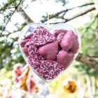 A transparent heart-shaped ornament filled with pink chocolate hearts is hanging from a tree branch. The chocolates are sprinkled with white dots. The background is blurred with hints of green foliage.