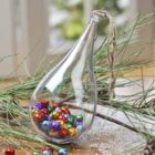 A teardrop-shaped glass ornament filled with colorful small jingle bells. It is resting on a table decorated with pine branches. A twine loop is attached at the top for hanging.