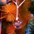 A delicate glass ornament shaped like a teardrop with a pink ribbon is hanging on a Christmas tree. The tree is decorated with shiny, orange tinsel, creating a festive and sparkling appearance.