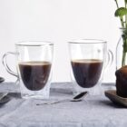 Two glass mugs of coffee on a table with a small milk bottle, a spoon, and forks on the left. A loaf of bread on a wooden board is on the right. In the background, a vase with green flowers adds a decorative touch.