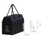 A pair of transparent glass mugs with handles sits next to a black gift box. The box has string handles and a simple design featuring a mug illustration and some text.