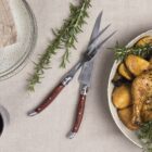 A roasted chicken garnished with herbs and lemon slices in a bowl, accompanied by a carving knife and fork on a linen tablecloth. Nearby are two sprigs of rosemary, a stack of plates with a napkin, and a glass of red wine.