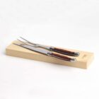 A carving set featuring a fork and a knife with wooden handles, resting on a light wooden box against a white background. The utensils have polished metal blades and tips, and the box has minimal text on one side.
