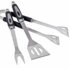 Three stainless steel BBQ tools are crossed: a fork, spatula, and tongs. Each has black and silver handles. The spatula and fork have cut-out designs on their blades. The tools are placed on a white background.