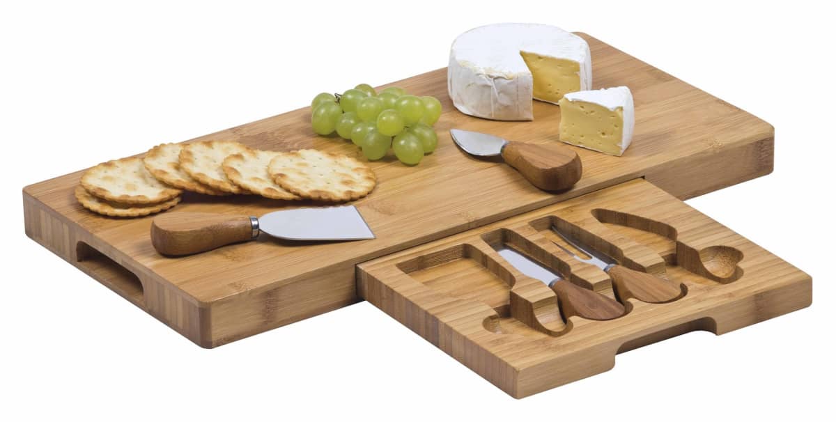 A bamboo cheese board set with tools and a built-in drawer containing four cheese knives. The board displays sliced cheese, green grapes, and round crackers. Another piece of cheese is partially sliced beside the arrangement.