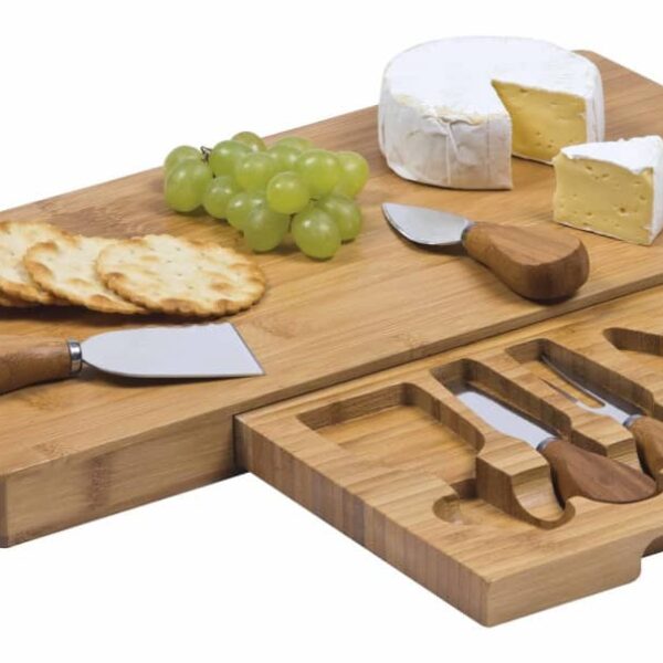 A two-tiered bamboo cheese board with a variety of cheese knives, crackers, green grapes, and a wheel of cheese with a slice cut out.