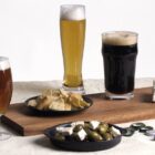 Three glasses of beer in different styles are on a table with small plates of crackers, olives, and cheese. Bottle caps and a bottle opener are nearby, set against a white background.