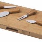 A rectangular bamboo cheese board with a drawer, featuring four cheese knives with wooden handles: a spreader, a serving fork, a flat chisel, and a pointed knife, neatly arranged on top.