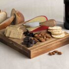 A wooden board with crackers, cheese, almonds, chocolate, and dried fruits. Sliced bread is in the background. A glass of red wine and a bottle partially visible. A cheese knife rests on the board.