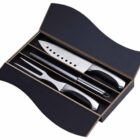 A black presentation box containing a stainless steel carving knife, carving fork, and sharpening steel, all with black handles. The box has a wavy lid design.