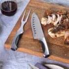 A wooden cutting board with a carving fork and knife beside sliced and whole pieces of roasted chicken. Two glasses of red wine and two empty plates are visible on the table, set with silverware and a gray napkin.
