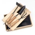 A wooden box with three gardening tools on top, including a trowel, a transplanter, and a hand rake. Each tool has a wooden handle and a leather strap loop. The box lid is slightly open.