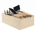 A light wooden box with three gardening tools on top: a trowel, a rake, and a spade. Each tool has a metal head and a light wood handle.