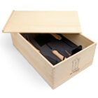 A wooden box with a sliding lid partially open, revealing gardening tools including a trowel, a hand rake, and a weeder inside. The box has a minimalist design with a small illustration on one side.