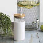 A glass beverage dispenser with a spigot, filled with water and lemon slices, sits on a rustic wooden surface. A white soap dispenser with a wooden top is next to a potted plant and fresh limes and a lemon.