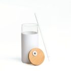 A minimalist white cup with a white silicone sleeve is accompanied by a white straw and a bamboo lid. The cup and its components are arranged on a plain white background.