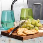 A wooden board with green grapes, crackers, dried apricots, a wedge of blue cheese, and a cheese knife. Two colored glasses, one teal and one green, are filled with water. Theres a metal chair and a light wooden background.