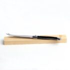 A stainless steel cheese knife with a black handle, featuring a two-pronged tip, is elegantly displayed on a light wooden block against a plain white background.