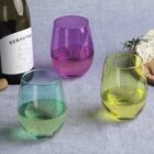 A table setting with a bottle of wine, three stemless wine glasses in blue, purple, and yellow, a cork, and a cheese platter with crackers and herbs on a light gray tablecloth.