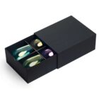 A black box partially opened to reveal four colorful glass tumblers inside. The tumblers are in shades of teal, amber, and violet, neatly arranged in separate compartments.