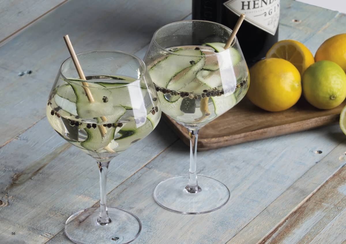 Two glasses of gin and tonic with cucumber slices and peppercorns on a wooden table. A bottle of Hendricks gin and a cutting board with lemons and limes are in the background.