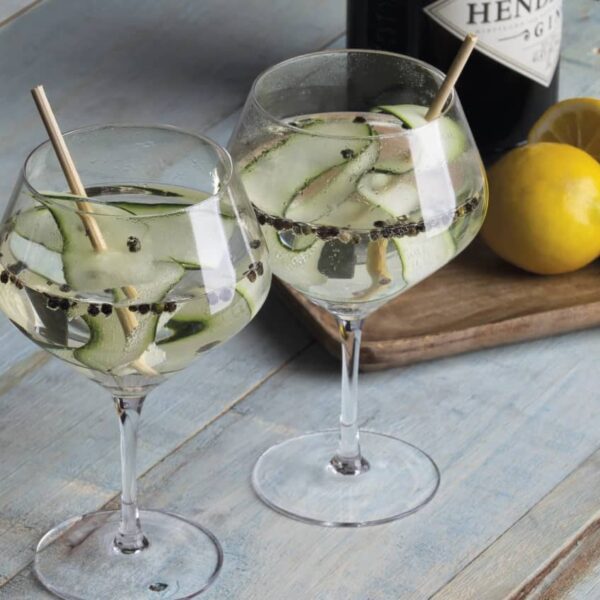 Two gin and tonics in balloon glasses with cucumber slices and floating peppercorns, on a rustic wooden table. A bottle of gin and a cutting board with lemons and limes are in the background.