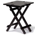 A small, black folding table with a simple design. The tabletop is divided into two sections and has holes for easy carrying. The legs form an X-shape for stability.