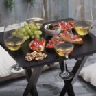 A small black table with four glasses of white wine, a cluster of green grapes, walnut pieces, and bruschetta topped with tomatoes and herbs. The setting includes two white cushions in a cozy, wooden-floored space.