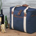 A navy blue cooler bag with beige straps sits on a wooden surface. Next to it are three wine bottles, a beer bottle, a jar of mixed dried fruit, and a jar with crackers. Greenery hangs in the background.
