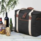 A black insulated cooler bag with beige straps sits on a wooden surface. Nearby are four bottles of various drinks, a mason jar filled with dried fruits, and wrapped food items. Green leaves hang from above, adding a touch of nature to the scene.