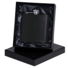 A black flask with a silver cap is nestled in a satin-lined black gift box. The lid of the box is propped underneath it.