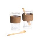 Two clear double-walled glass cups with brown cork bands around the middle. One cup contains a wooden spoon, and another identical wooden spoon lies in front of them. The cups are clean and placed against a plain white background.