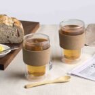 Two glasses of tea with cork sleeves on a table set with sliced bread, butter, a wooden spoon, and a newspaper with a crossword puzzle. A beige cloth napkin is draped in the background.